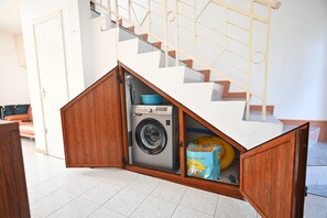 A compact laundry area with a washing machine and a blue bag inside a wooden cabinet under a staircase.
