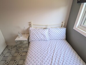 A neatly made bed with floral-patterned bedding, a bedside table with a decorative item, and a patterned floor. - 23 Cherry Park (Skegness)