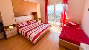 A bedroom with a large bed, a smaller bed, wooden cabinets, a nightstand, and a window with a view. - Apartman Kira 4 (Dramalj)
