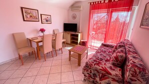 A living room with a sofa, a TV, a dining table with chairs, and a window with red curtains. - Apartman Kira 4 (Dramalj)