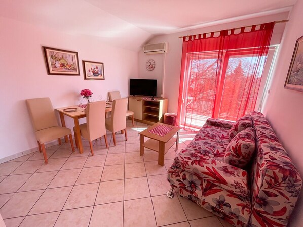 Un salon comprenant un canapé, un téléviseur, une table à manger avec des chaises et une fenêtre avec des rideaux rouges.
