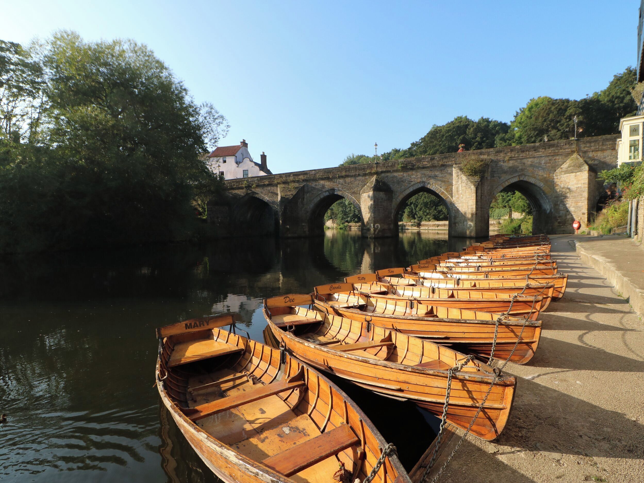 A row of wooden boats docked by a river with a stone bridge in the background.