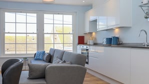 A modern kitchen with white cabinets, a built-in oven, and a microwave. There is a grey sofa with cushions in the living area, and a window with a view of a yellow building. - The Seven Sands (Romney Marsh)