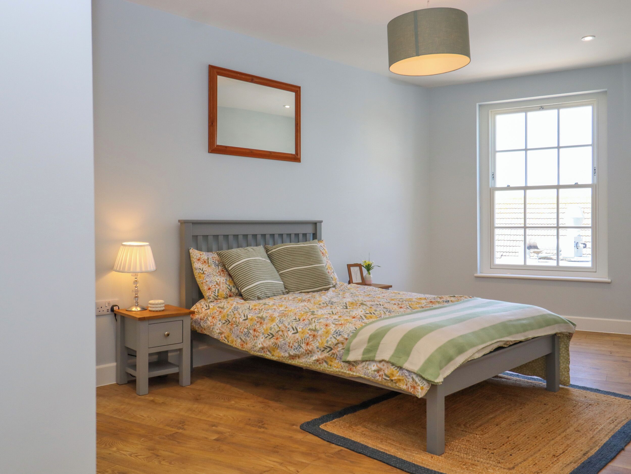 A bedroom with a bed, a nightstand, a lamp, a mirror, and a window.