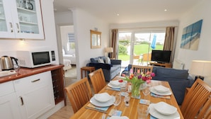 A dining area with a wooden table set for four, surrounded by chairs. A kitchen area with a microwave and cabinets is visible to the left. - Cape A1 (Yarmouth)