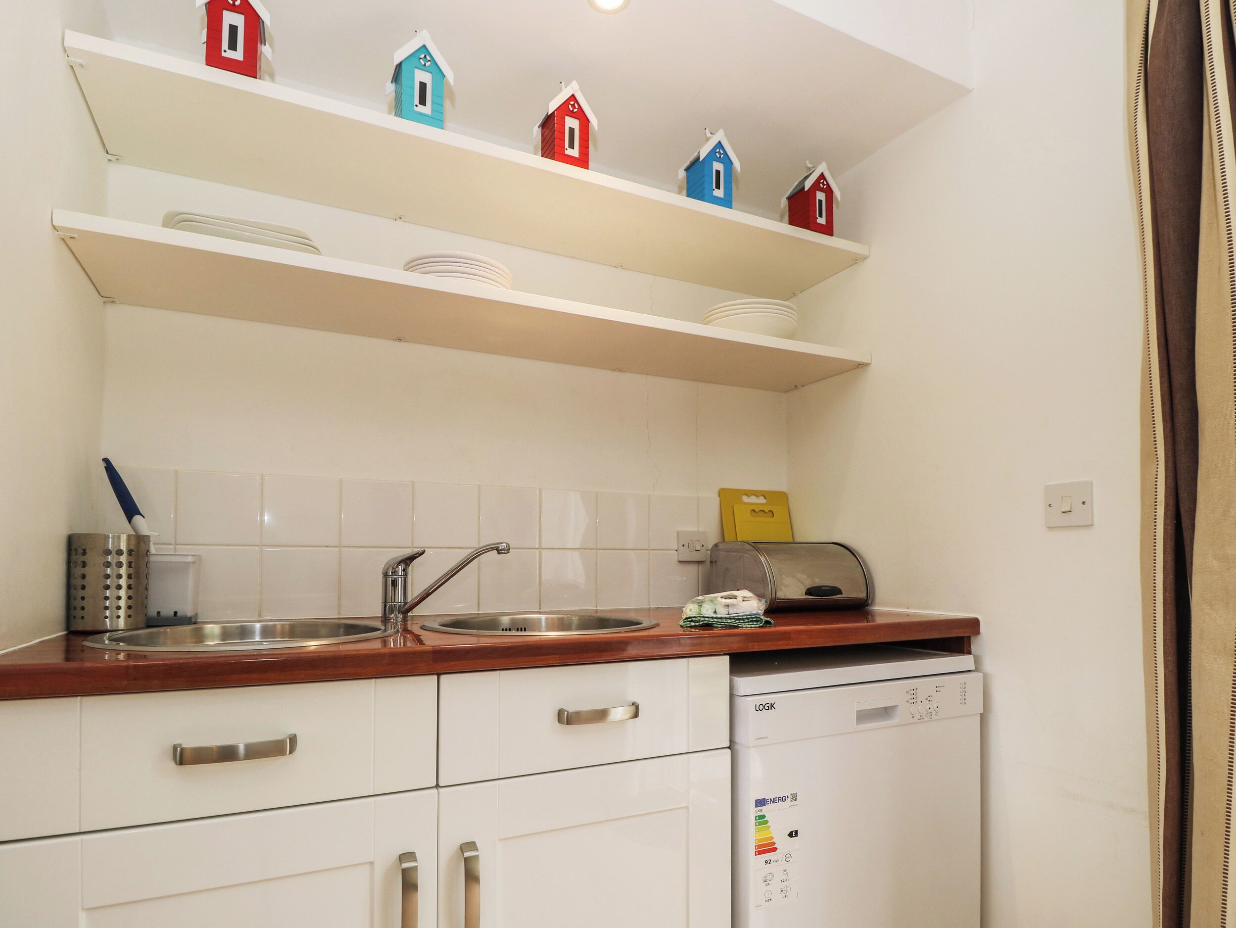 A kitchen with white cabinets, a stainless steel sink, and a dishwasher. There are colorful house-shaped decorations on the shelves.