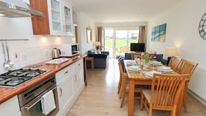 A modern kitchen with a gas stove, oven, and microwave. There is a dining area with wooden chairs and a table set for four. A TV is mounted on the wall, and a window provides a view of the outdoors. - Cape A1 (Yarmouth)