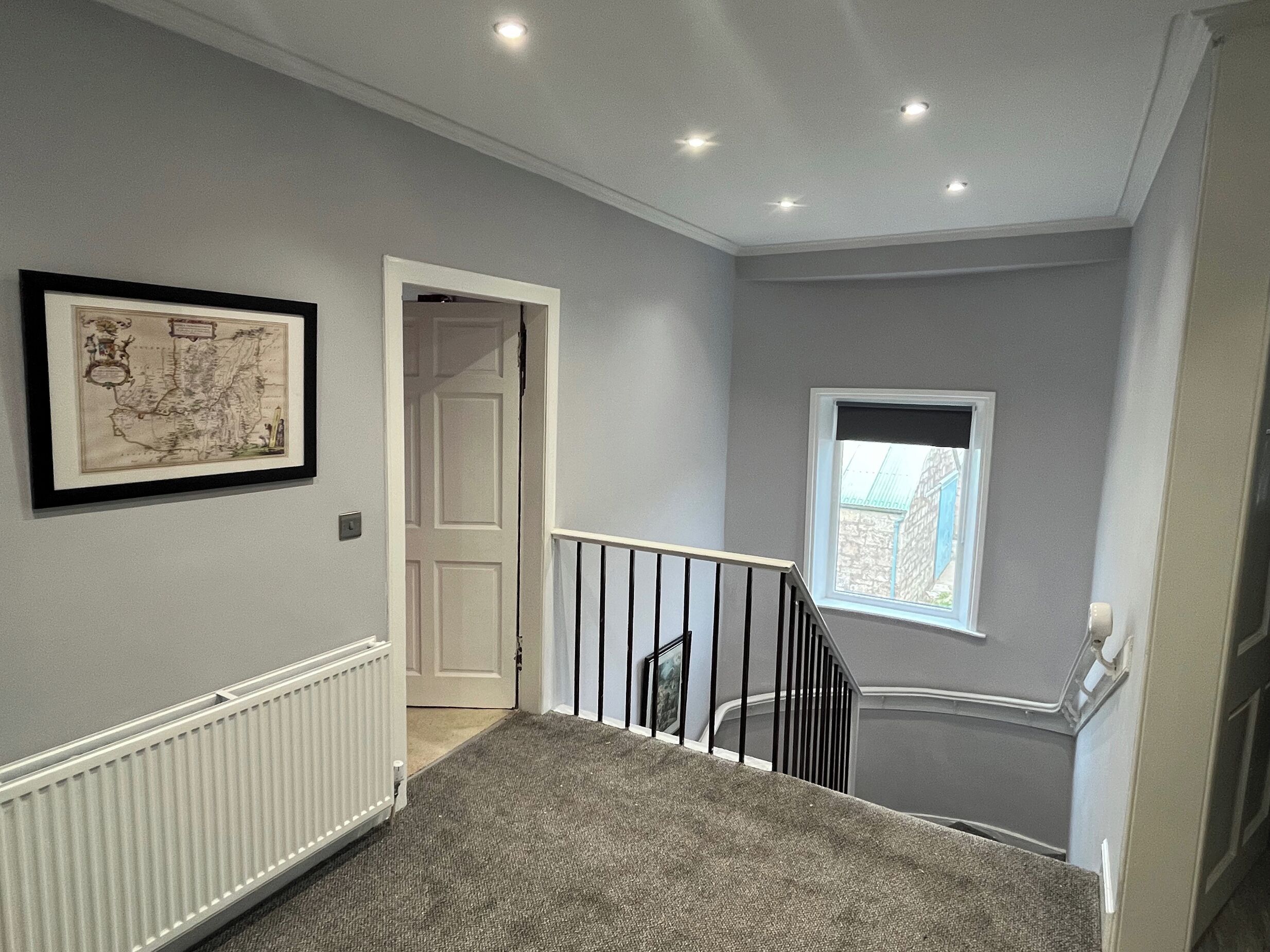 A hallway with a radiator, a framed map, a door, a staircase with a black railing, and a window with a view of the outside.