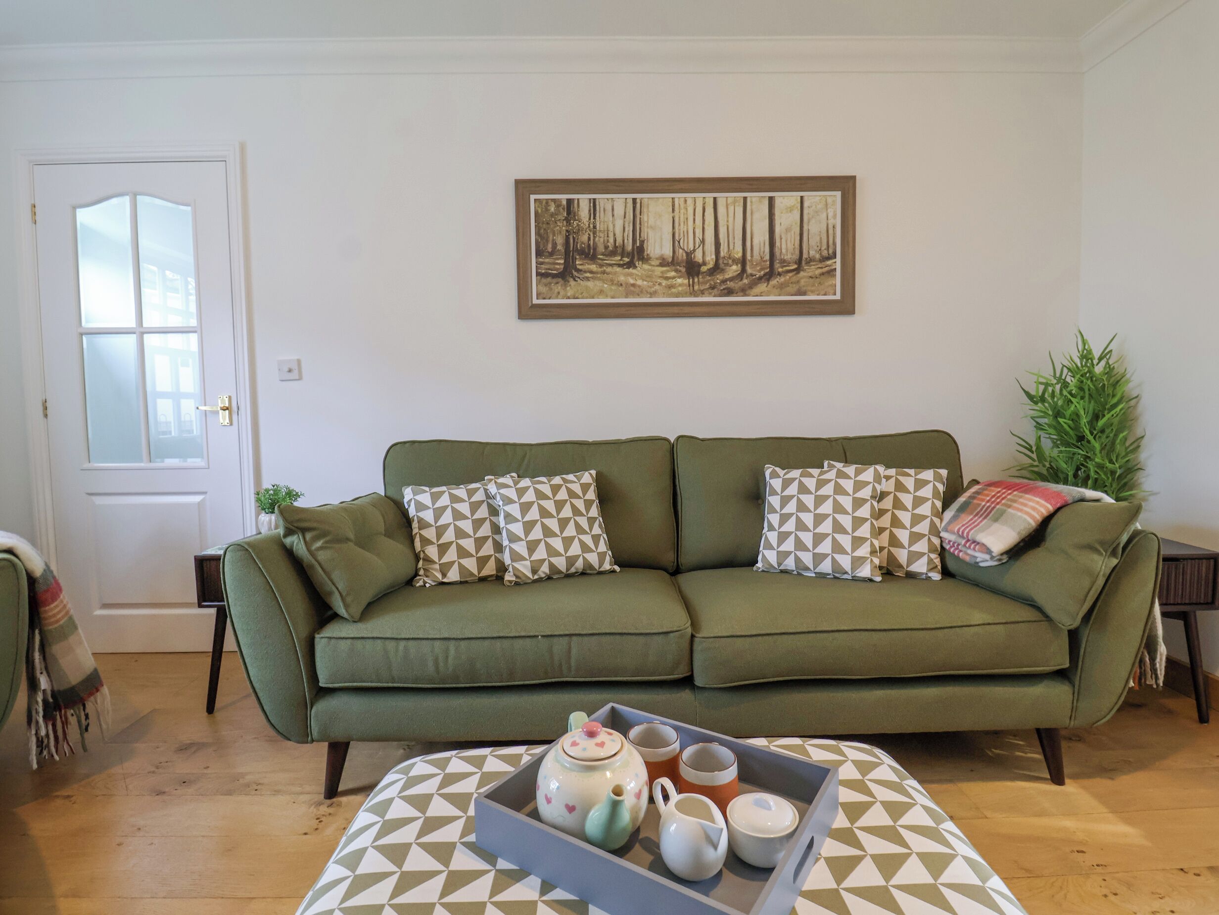 Un soggiorno con un divano verde, cuscini decorati, un vassoio con teiere e tazze, e un quadro raffigurante una foresta.