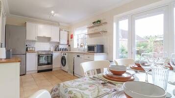 A compact kitchen with a washing machine, oven, and microwave. There is a dining area with a table set for four, and a window providing a view of the outside.