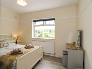 A bedroom with a bed, a window, a radiator, a bedside table with a lamp, and a mirror on a dresser. - Greenacres (Kidderminster)