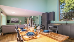 Comedor con mesa de madera dispuesta para seis personas, rodeada de sillas, con vistas a la cocina y una escalera que sube.