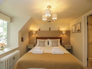 A bedroom with a large bed, two bedside tables, a radiator, a window, and a chandelier. - Thorn Cottage (Fairford)