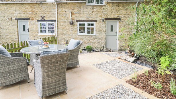 A stone house with a tiled patio, wicker furniture, and a small garden area. - Thorn Cottage (Fairford)
