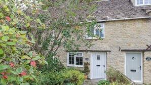 A stone cottage with a white door, a small window, and a garden with various plants and a gravel path. - Thorn Cottage (Fairford)