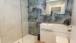 Een moderne badkamer met een douche met glazen wand, een marmeren spatwand, een witte wastafel en een toilet.