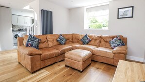 A modern living room with a brown leather sectional sofa, a wooden coffee table, and a kitchen visible in the background. - Fern Cottage (Stoke-on-Trent)