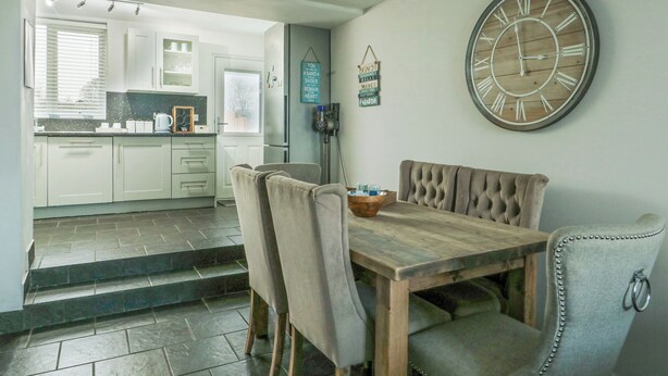 Un comedor con una mesa y sillas de madera, un reloj grande en la pared y vistas a la cocina.
