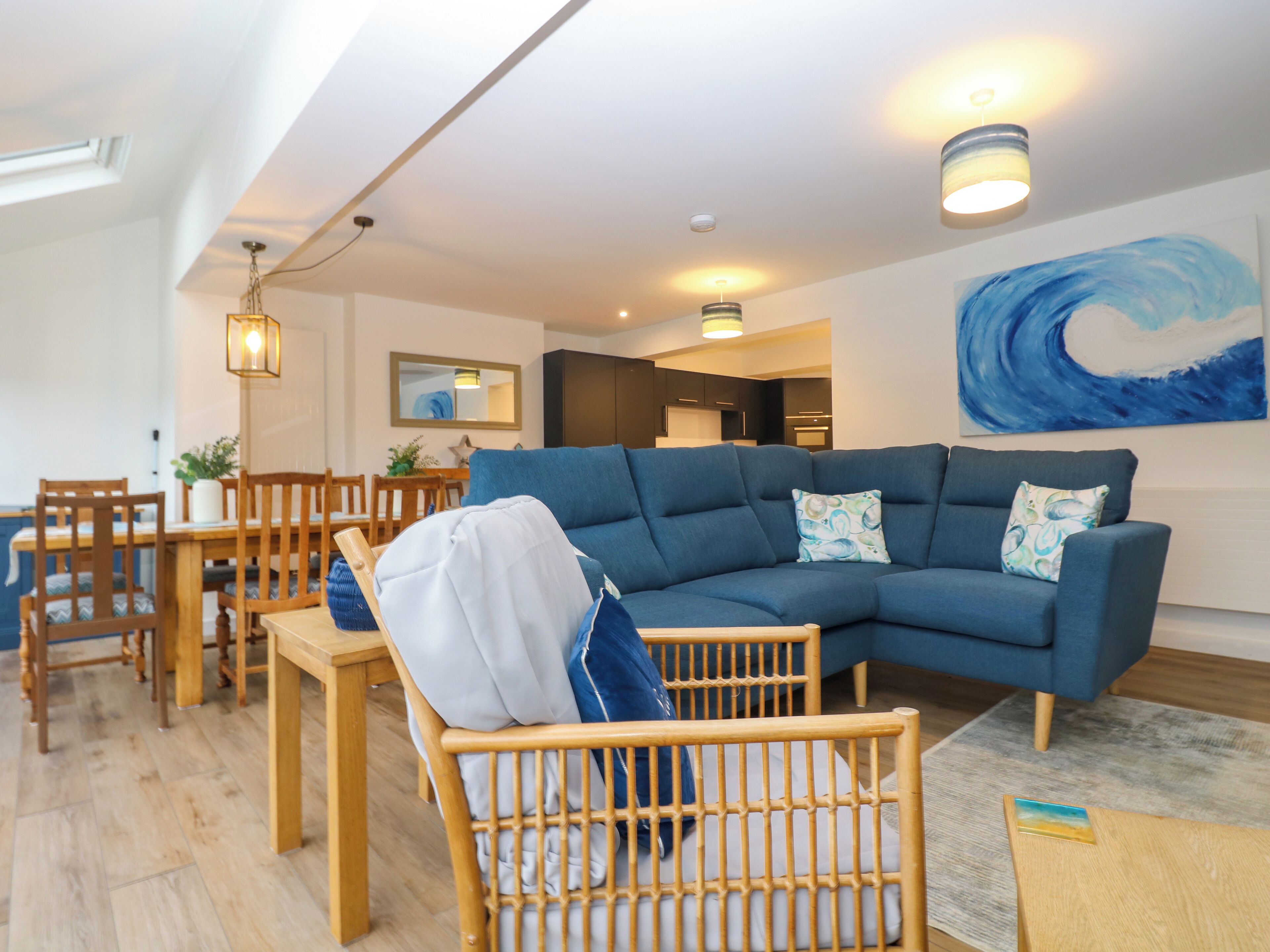 Un salon moderne avec un canapé d’angle, une table à manger en bois et des chaises. Un grand tableau orne le mur et un luminaire est fixé au plafond.