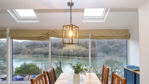 A dining area with a large table, chairs, and a view of a river and forest through large windows. - Anjoma (Looe)