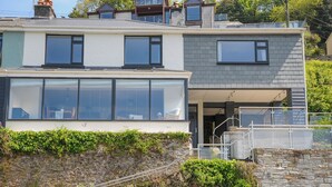 A modern two-story house with large windows, a stone wall, and a balcony. - Anjoma (Looe)