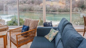 A living room with a blue sofa, a wicker chair, and a view of a lake through large windows. - Anjoma (Looe)