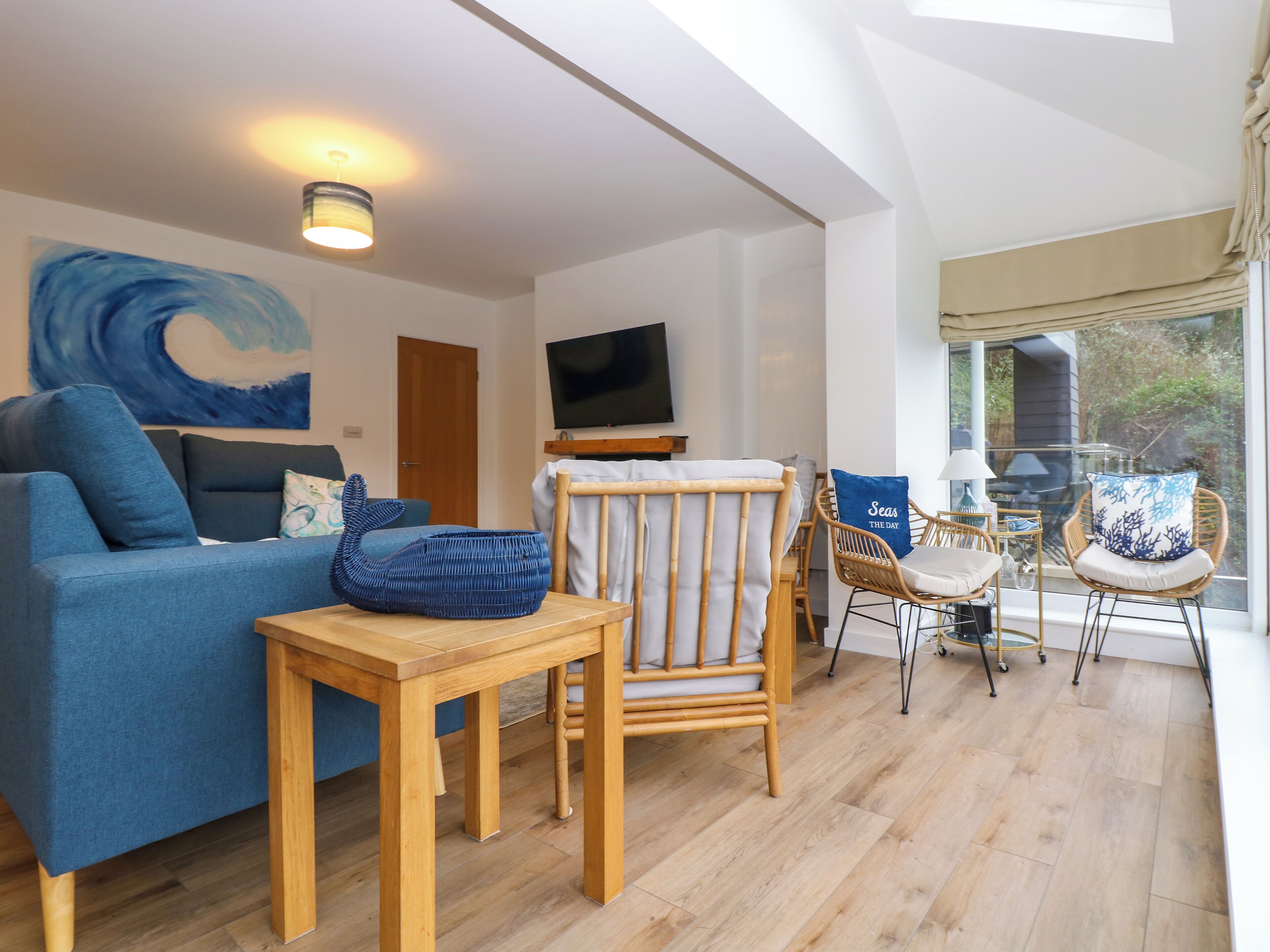 A living room with a blue sofa, a wooden table, wicker chairs, and a TV on the wall.