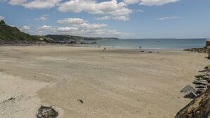 A wide sandy beach with a rocky shoreline, a clear blue sky, and distant hills. - Anjoma (Looe)