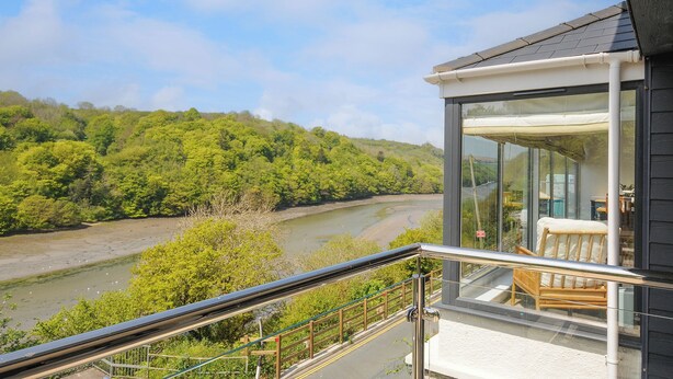 A modern building with a glass balcony overlooking a river and forested hills. - Anjoma (Looe)