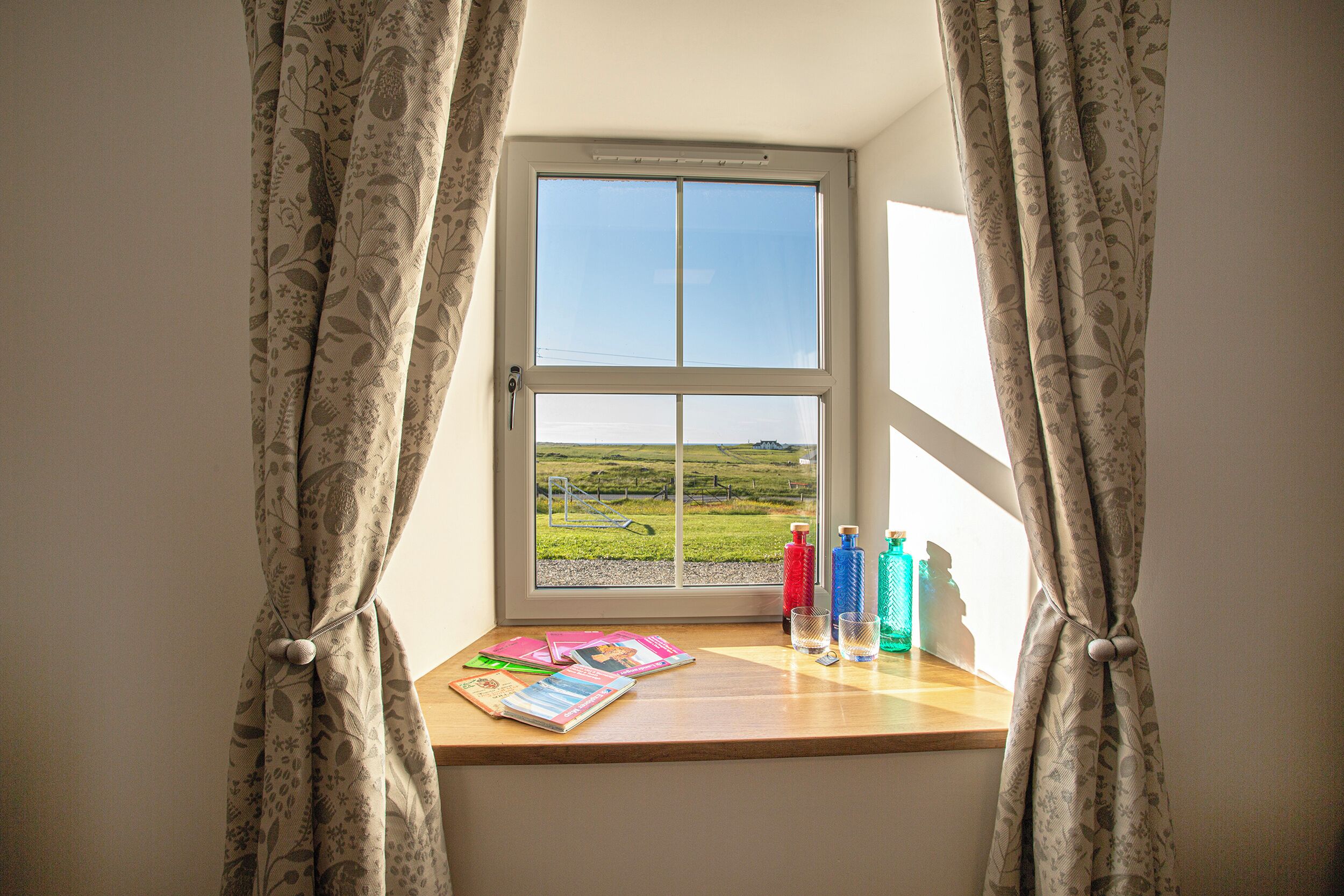 A window with curtains tied at the top, a wooden ledge with magazines, and two bottles on the windowsill.