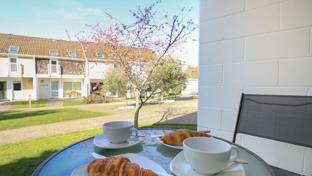 Ett bord med ett croissant, en kopp och en liten assiett på en balkong med utsikt över en trädgård och byggnader.