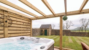 Un jacuzzi bajo una pérgola de madera, con un muro de piedra y un edificio de piedra al fondo.