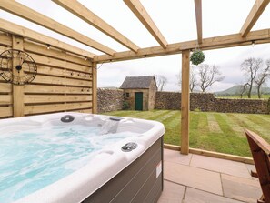 Un bain à remous sous une pergola en bois, avec un mur de pierre et un bâtiment en pierre en arrière-plan.