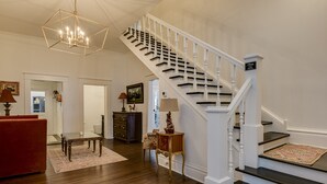 A spacious living room with a red sofa, a wooden coffee table, and a chandelier. A staircase with white railings leads to another level. - Burrell and Flynn's Farm (Aiken)