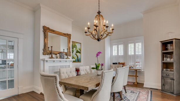 A dining room with a wooden table, white upholstered chairs, a chandelier, a large mirror, and a patterned rug. - Burrell and Flynn's Farm (Aiken)