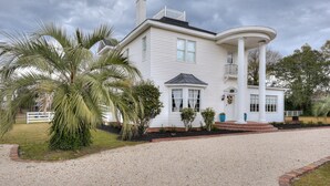 A white two-story house with a curved front porch, a chimney, and a gravel driveway. - Burrell and Flynn's Farm (Aiken)