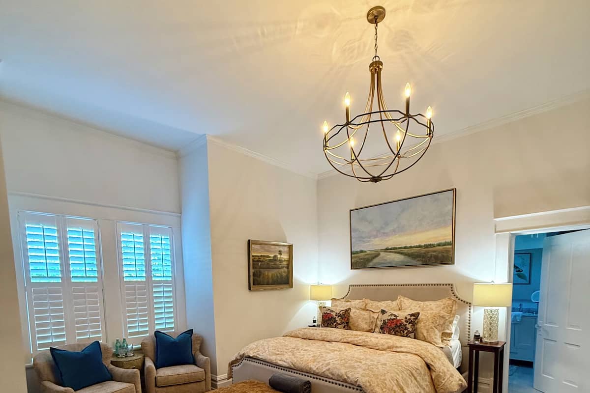 A bedroom with a large bed, two armchairs, a side table, a chandelier, and a painting on the wall.