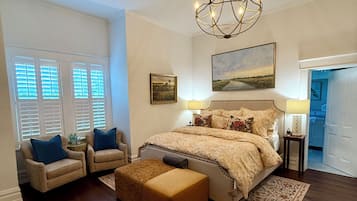 A bedroom with a large bed, two armchairs, a side table, a chandelier, and a painting on the wall.