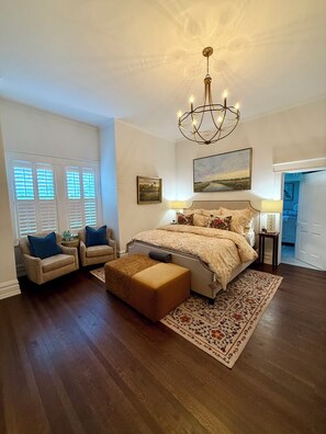 Une chambre à coucher avec un grand lit, deux fauteuils, une table d’appoint, un lustre et un tableau au mur.