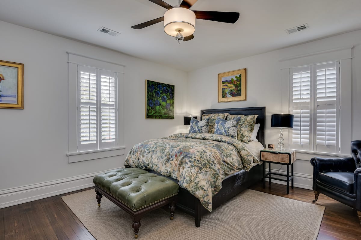 A bedroom with a bed, a ceiling fan, a nightstand, a chair, and artwork on the walls.