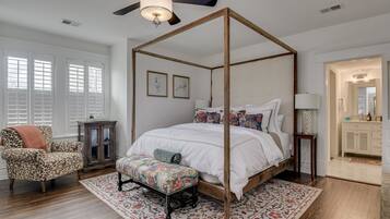 A four-poster canopy bed with a white bedspread and colorful pillows, a wooden chair with a patterned cushion, a small cabinet, and a patterned rug.