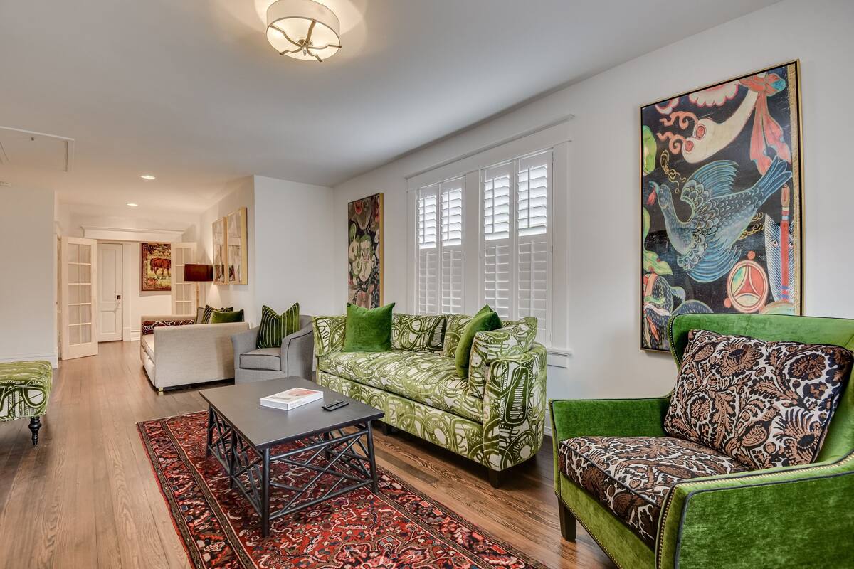 A modern living room with a green patterned sofa, a matching armchair, and a coffee table. There is a large painting on the wall and a window with shutters.