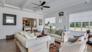 A spacious living room with a high ceiling, a ceiling fan, and a large window with a view of the outside. There are two sofas and a chair, a coffee table, and a rug. - Burrell and Flynn's Farm (Aiken)