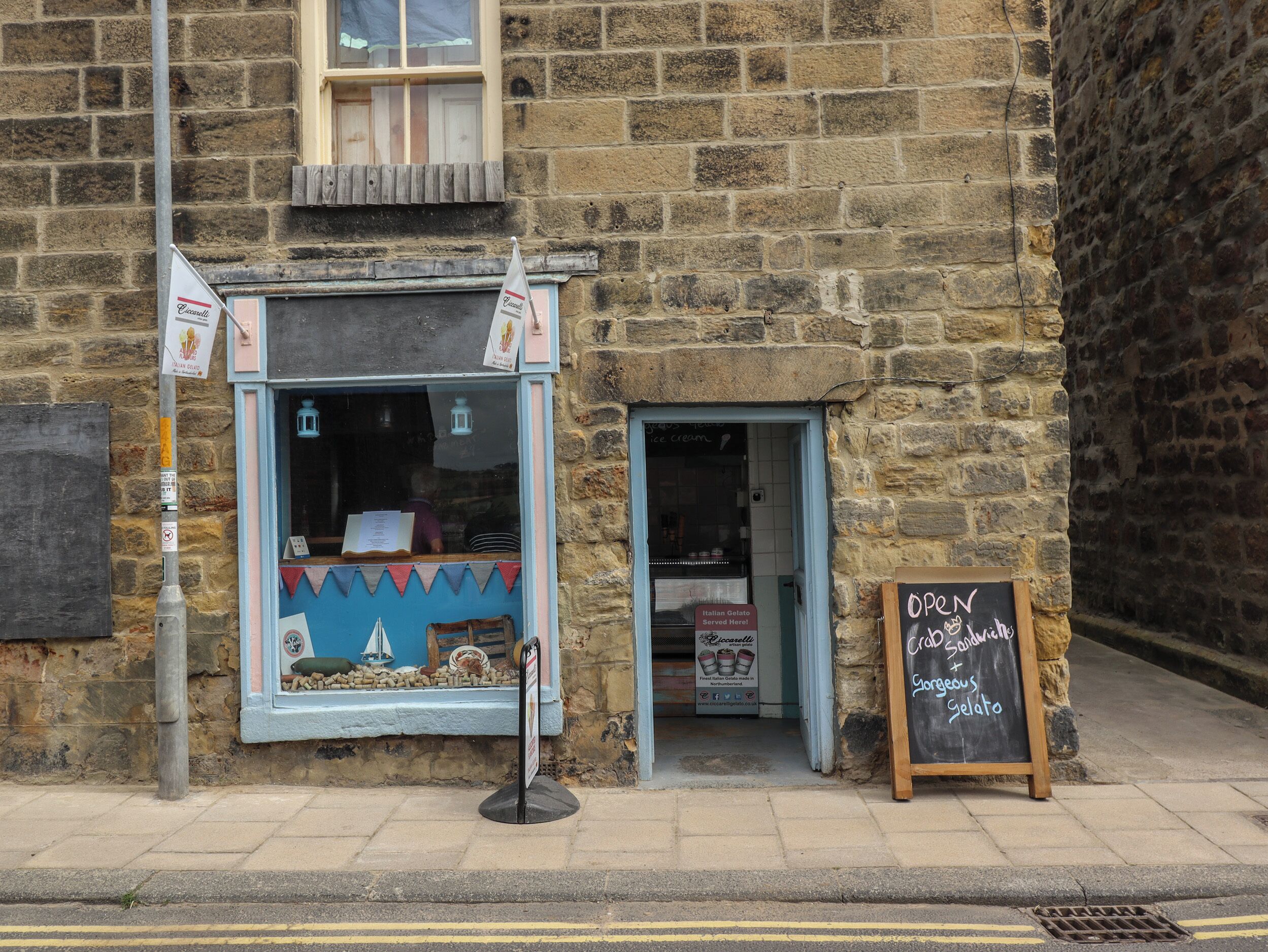 A stone building with a small shop entrance, a signboard for Italian Gelato, and a chalkboard sign advertising open hours.