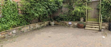 A paved courtyard with a brick floor, surrounded by a stone wall and dense foliage.