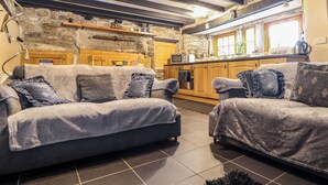 A cozy living room with a stone wall and wooden beams, a kitchen with wooden cabinets, and a large window. - Up Over Head (Penzance)