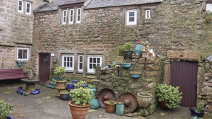 A stone building with multiple windows and doors, surrounded by various potted plants and outdoor furniture. - Up Over Head (Penzance)