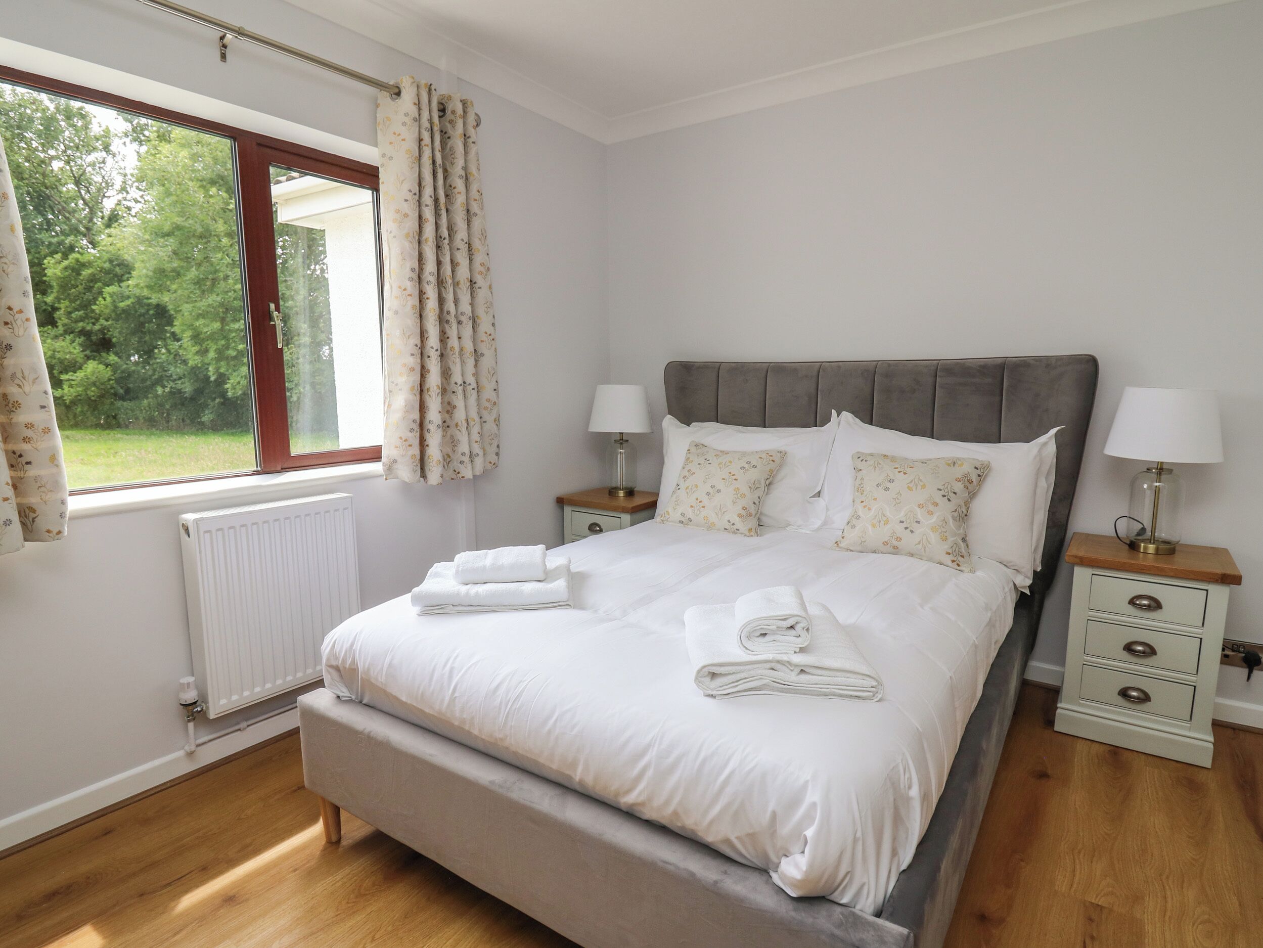 A neatly made bed with pillows and a folded towel, a bedside table with a lamp, and a window with curtains.