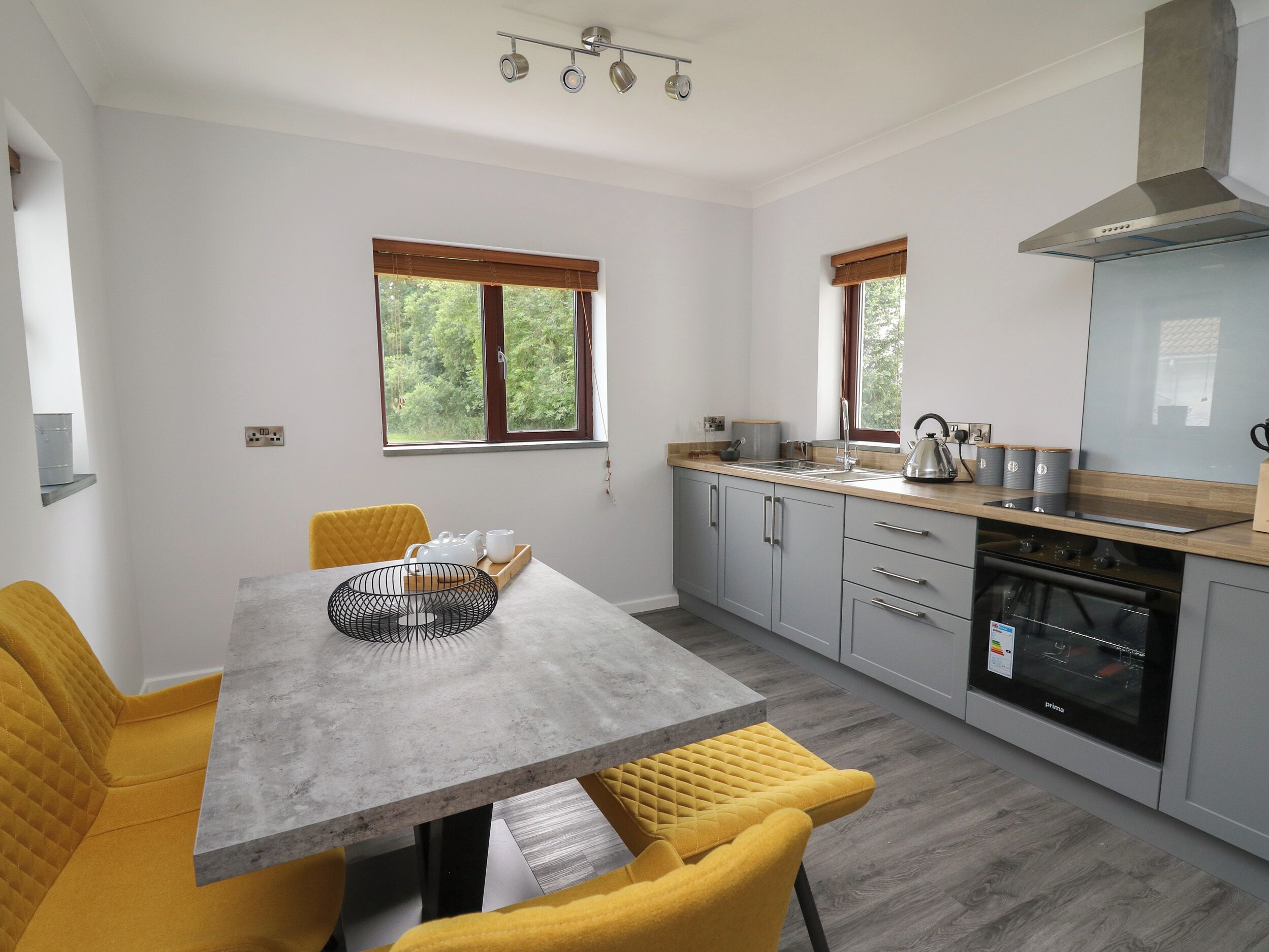 A modern kitchen with grey cabinets, a wooden countertop, and a built-in oven. There are two windows with wooden blinds, a dining table with yellow chairs, and a kettle on the counter.