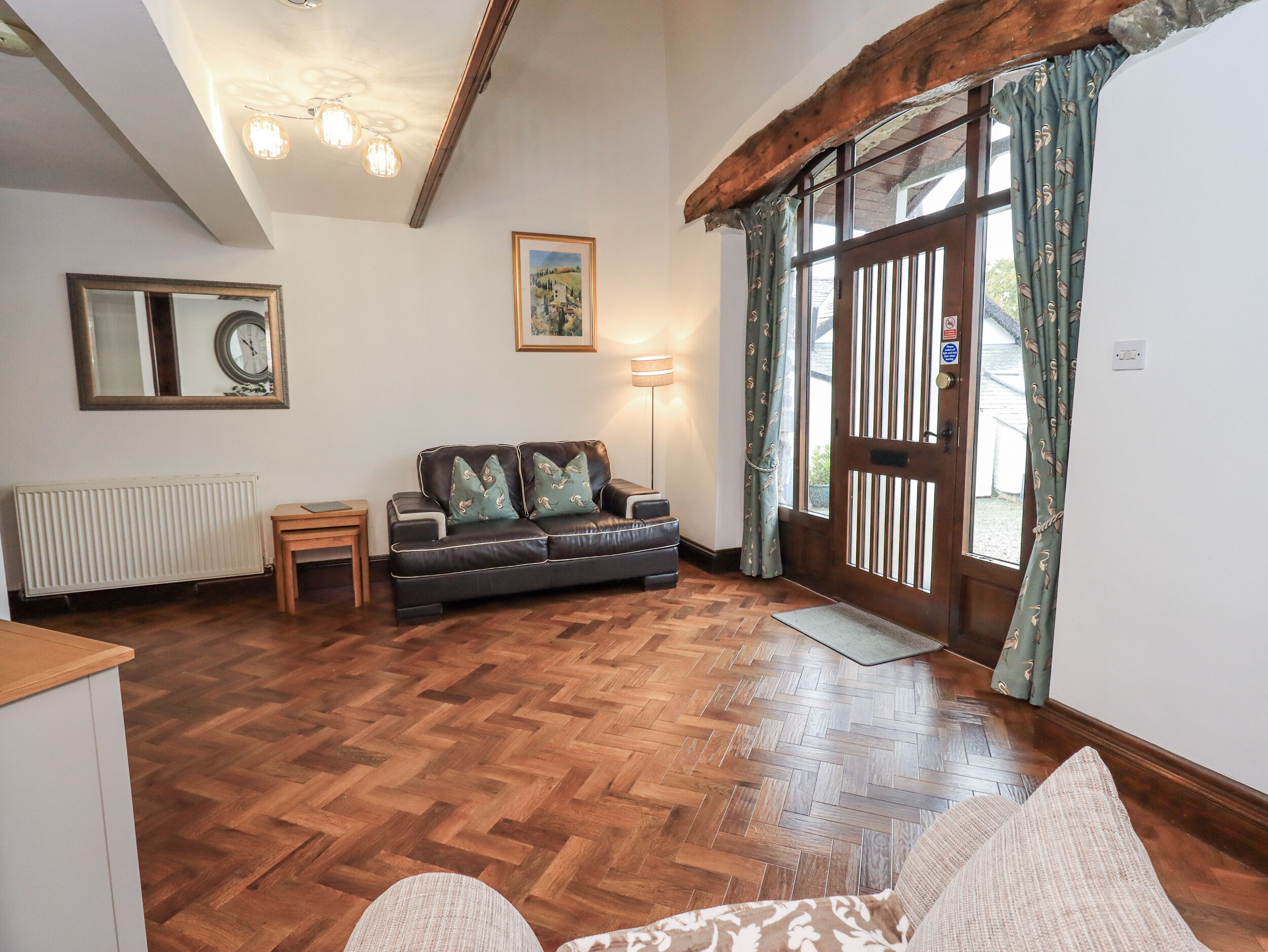 A living room with a herringbone wooden floor, a dark leather sofa with patterned cushions, a wooden side table, a framed picture on the wall, and a large window with curtains.